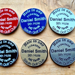 May include: Six golf ball markers, each with "Hole in One", the date, name "Daniel Smith", hole details, and golf club name. The markers are in various colours: red, blue, white, black, gold, and silver. Yardage is given in yards.