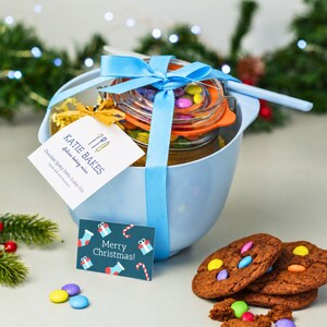 May include: A light blue mixing bowl containing a glass jar filled with cookie mix and colourful sweets, tied with a blue ribbon. A tag reads Katie Bakes Artisan baking mixes Chocolate Spotty Dotty Cookie Mix and a card says Merry Christmas.