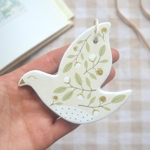 Puede incluir: Adorno en forma de paloma de color crema con un diseño floral en verde y dorado. El adorno tiene una cuerda para colgar. El ala de la paloma tiene un patrón de puntos. El adorno se sostiene en una mano.