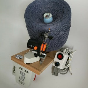 May include: A gray yarn ball is on a wooden base with a white electric yarn winder. The winder has a digital counter and an orange knob. A white control box with a red button and a knob is connected to the winder.