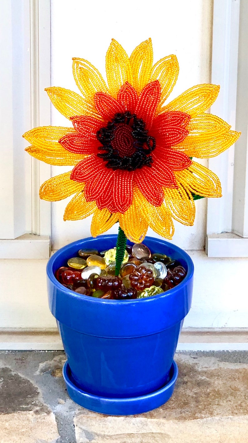 May include: A beaded sunflower with yellow and orange petals and a black center is in a blue ceramic pot with glass stones.