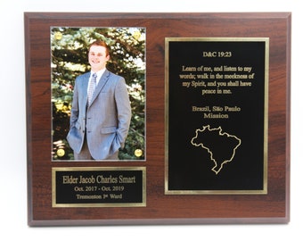 Puede incluir: Una placa de madera con un borde dorado y un fondo negro. La placa tiene una foto de un joven en traje, una inscripción dorada con el texto "Elder Jacob Charles Smart Oct. 2017-Oct. 2019 Tremonton 1st Ward", y una inscripción dorada con el texto "D&C 19:23 Aprended de mí, y escuchad mis palabras; andad en la mansedumbre de mi Espíritu, y tendréis paz en mí. Brasil, São Paulo Mission".