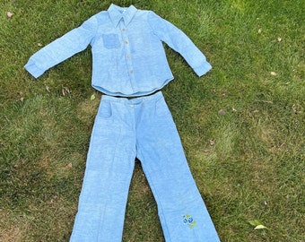 May include: A light blue two-piece outfit, consisting of a button-down shirt and flared pants. The pants feature a floral embroidery near the hem. The outfit is made of a textured fabric and is laid out on green grass.