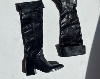 May include: A pair of black leather over-the-knee boots with a block heel. The boots have a fold-over cuff at the top and a zipper on the inside of the shaft.