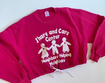 May include: A pink sweatshirt with a white graphic that says "Share and Care Center Neighbors Helping Neighbors". The sweatshirt has a white drawstring at the bottom.