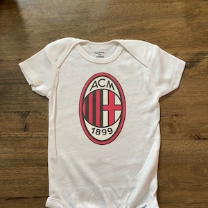 May include: White baby onesie with the AC Milan team logo printed on the front. The logo is an oval shape with the letters "ACM" at the top, a red and black striped section, and the year "1899" at the bottom.