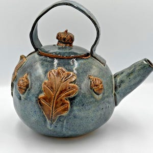 May include: A ceramic teapot with a blue-green glaze. The teapot features a large brown oak leaf and acorn designs. The handle and spout are integrated into the design. The lid is topped with a small acorn.