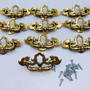 May include: A set of ten brass-colored metal drawer pulls featuring a decorative, ornate backplate with a curved, swinging handle. Each pull has a polished finish and is accompanied by a collection of small, silver-colored metal mounting screws.