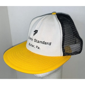 Può includere: Cappellino da camionista con pannello frontale bianco con la scritta "Hillman Standard Butler, Pa." e un logo nero. La visiera è gialla e il retro in rete è nero. Un bottone giallo è in alto.