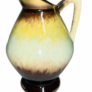 May include: A small, ceramic pitcher with a brown, yellow, and white glaze. The pitcher has a rounded body and a flared lip. The handle is attached to the side of the pitcher and curves up and over the top.