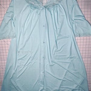 May include: A light blue duster robe featuring a V-neckline with white lace trim, short sleeves with lace cuffs, a single patch pocket, and a front button closure. The garment is displayed against a ruler showing a width of 31 inches.