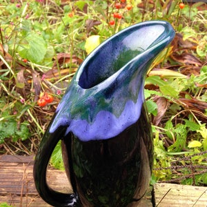 May include: A handcrafted ceramic pitcher with a glossy finish. The pitcher features a black base, transitioning to a vibrant purple and green rim. The handle is curved, and the spout is elongated. The pitcher is set against a natural outdoor backdrop.