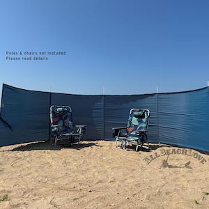 May include: A navy blue beach windbreak with white poles, set up on a sandy beach. Two folding beach chairs with a floral pattern are positioned in front of the windbreak. The sky is clear and blue.