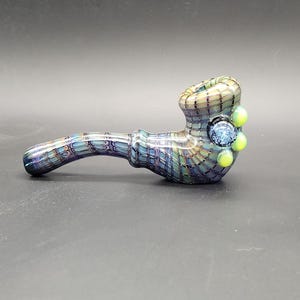 May include: A hand-blown glass pipe with a textured, grid-like pattern in shades of blue, green, and purple. The bowl features a dark blue center with light green accents and three small, round, light green embellishments.
