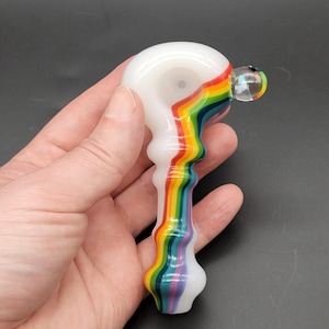 May include: A hand-held glass pipe featuring a smooth, opaque white finish with a vibrant, multi-colored rainbow stripe pattern running vertically along the stem and bowl. The pipe has a rounded bowl, a textured stem, and a small clear glass carb.