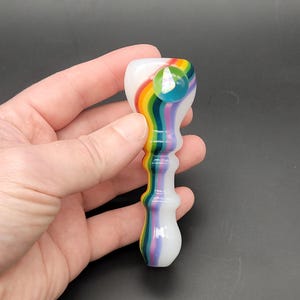 May include: A glass pipe featuring a white base with vertical rainbow stripes in red, orange, yellow, green, blue, and purple. The top section includes a clear, circular glass marble accent with an iridescent, reflective, and multi-colored finish.