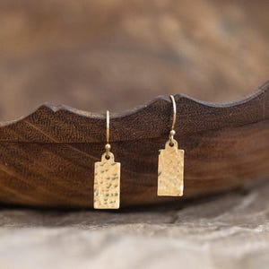Könnte beinhalten: Ein Paar goldfarbene, rechteckige Ohrhänger mit gehämmerten Strukturen. Die Ohrringe hängen an kleinen, gebogenen Haken. Der Hintergrund ist eine verschwommene Holzschale.