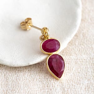 Op de afbeelding: Een enkele gouden oorbel met twee dieprode edelstenen. De bovenste steen is ovaal, de onderste druppelvormig. De edelstenen zijn gezet in gouden zettingen. Elegant ontwerp, op een witte achtergrond.