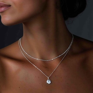 Könnte beinhalten: Eine silberne Kettenkette mit einem kleinen, gehämmerten Silberanhänger. Die Kette ist mit einer dünneren Silberkette geschichtet.
