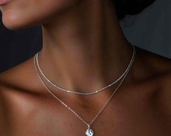 Op de afbeelding: Een gelaagde zilveren kettingenset bestaande uit een korte, delicate ketting met kleine, uniforme schakels en een langere, fijnere ketting met een enkele, kleine, ronde, gefacetteerde hanger die het licht helder en sprankelend weerkaatst.