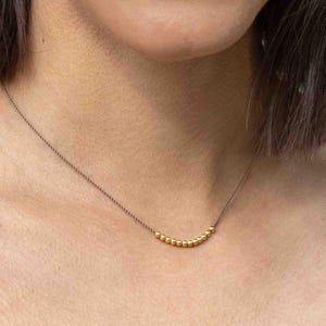 Könnte beinhalten: Eine zarte Halskette mit kleinen, goldfarbenen Perlen, die auf einer dünnen, braunen Kordel aufgereiht sind. Die Perlen sind in der Mitte angeordnet und bilden eine subtile, elegante Kurve. Die Halskette wird vor einem Hautton-Hintergrund gezeigt.