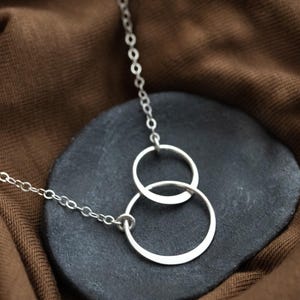 Könnte beinhalten: Eine silberne Halskette mit zwei ineinandergreifenden Kreisen, die an einer feinen Kette hängt. Der Anhänger liegt auf einer dunklen, strukturierten Oberfläche, vor einem braunen Stoffhintergrund. Das Design ist minimalistisch und modern.
