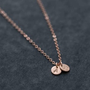 Op de afbeelding: Een delicate gouden ketting met twee kleine ronde hangers, elk gegraveerd met een letter. De letters zijn "A" en "M".