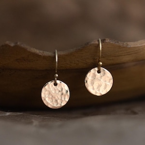 Könnte beinhalten: Ein Paar Ohrhänger mit kleinen, kreisförmigen, gehämmerten Metallscheiben in einem warmen Roségoldton. Jede Scheibe ist an einem einfachen, dünnen Metallhaken befestigt, was eine zarte und reflektierende Oberflächenstruktur auf den Anhängern erzeugt.