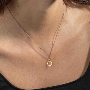 May include: A delicate gold necklace featuring a small, open circle pendant. The pendant hangs from a thin, brown chain. The necklace is worn against a person's skin, with a black top visible at the bottom of the frame.