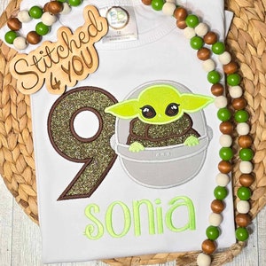 Op de afbeelding: Een witte romper met een groen, bruin en gouden glittercijfer negen, een groen personage uit de Star Wars-franchise en de naam "sonia" geborduurd in groen. Op de achtergrond is een houten bord met de tekst "Stitched 4 You" te zien.