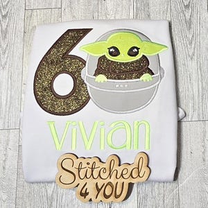 Op de afbeelding: Een wit shirt met een gouden glittercijfer zes, een groen personage uit Star Wars en de naam "vivian" geborduurd in groen. Een houten bord met de tekst "Stitched 4 YOU" bevindt zich in de rechterbenedenhoek.