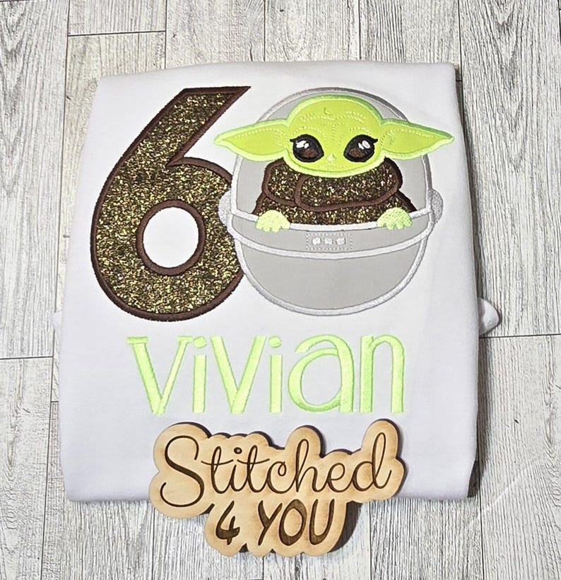 Op de afbeelding: Een wit shirt met een gouden glittercijfer zes, een groen personage uit Star Wars en de naam "vivian" geborduurd in groen. Een houten bord met de tekst "Stitched 4 YOU" bevindt zich in de rechterbenedenhoek.