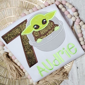 Op de afbeelding: Een wit T-shirt met een bruin en gouden glitter nummer 7, een groen geborduurd personage uit Star Wars en de naam "Audrie" geborduurd in groen.