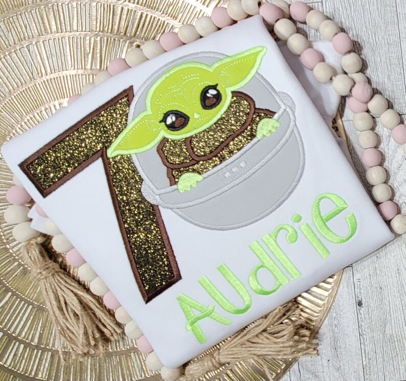 Op de afbeelding: Een wit T-shirt met een bruin en gouden glitter nummer 7, een groen geborduurd personage uit Star Wars en de naam "Audrie" geborduurd in groen.