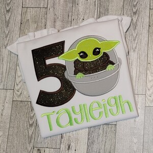 Op de afbeelding: Wit T-shirt met een geborduurd beeld van een personage uit Star Wars in groen, bruin en grijs. Het getal 50 is geborduurd in bruin en zwart glitter. De naam "Tayleigh" is geborduurd in groen.