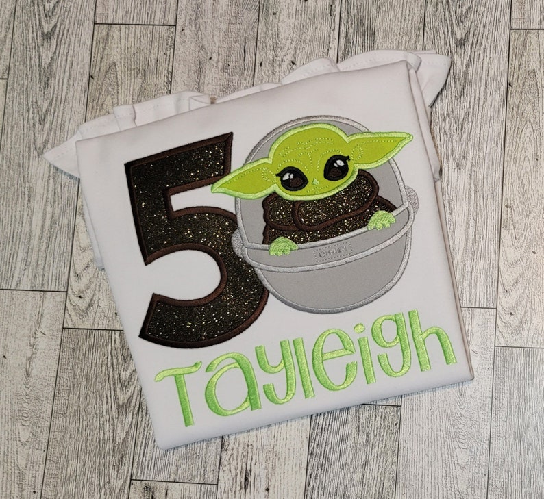 Op de afbeelding: Wit T-shirt met een geborduurd beeld van een personage uit Star Wars in groen, bruin en grijs. Het getal 50 is geborduurd in bruin en zwart glitter. De naam "Tayleigh" is geborduurd in groen.