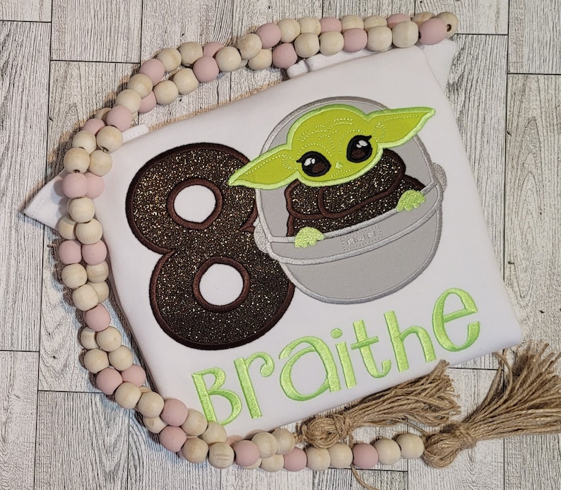 Op de afbeelding: Een wit T-shirt met een bruin glitterend nummer 8, een groen en grijs geborduurd personage en de naam "Braithe" in groene borduurwerk.
