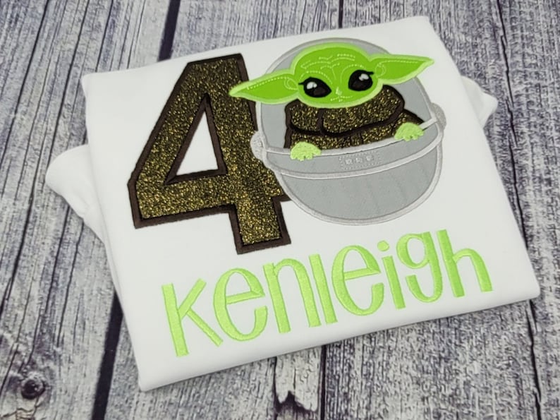 Op de afbeelding: Wit T-shirt met een gouden glitter nummer 40, een groen alien personage in een ruimteschip en de naam "kenleigh" in groen.