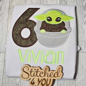 Op de afbeelding: Een wit T-shirt met een bruin en gouden glittercijfer 6, een groen alienfiguur in een grijze pod en de naam "Vivian" geborduurd in groen. Een houten bord met de tekst "Stitched 4 You" bevindt zich in de rechterbenedenhoek.