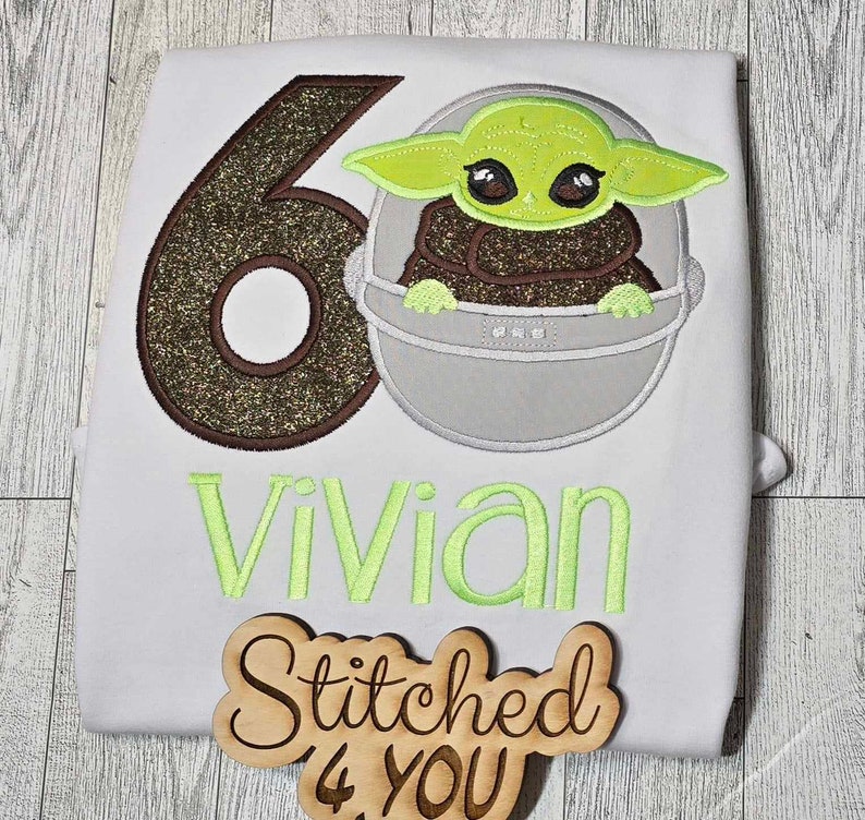 Op de afbeelding: Een wit T-shirt met een bruin en gouden glittercijfer 6, een groen alienfiguur in een grijze pod en de naam "Vivian" geborduurd in groen. Een houten bord met de tekst "Stitched 4 You" bevindt zich in de rechterbenedenhoek.