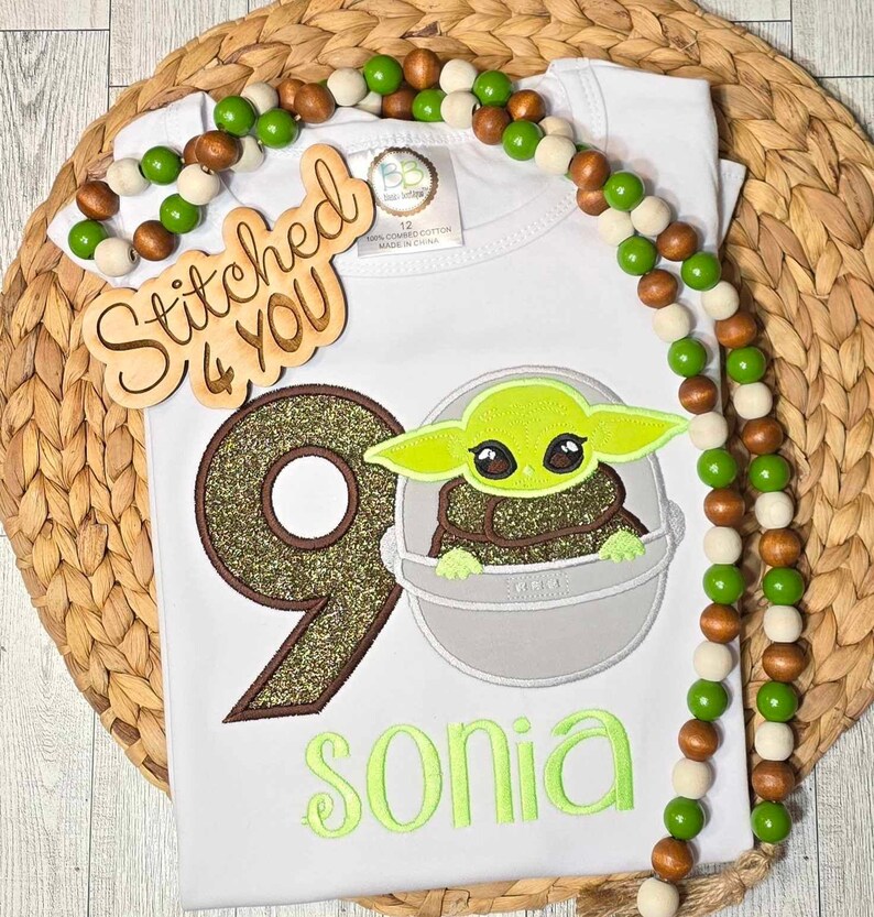 Op de afbeelding: Een witte romper met een groen, bruin en gouden glittercijfer negen, een groen personage uit de Star Wars-franchise en de naam "sonia" geborduurd in groen. Op de achtergrond is een houten bord met de tekst "Stitched 4 You" te zien.