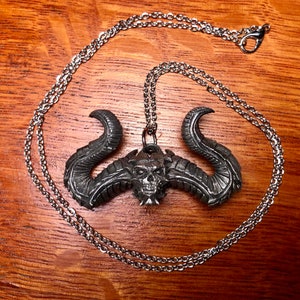 May include: A silver necklace with a detailed pendant of a demon head with horns. The pendant is made of metal and has a dark, aged finish.