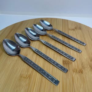 May include: Set of five silver spoons with an ornate floral design on the handles. The spoons are arranged on a wooden surface.