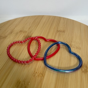 May include: Three heart-shaped bracelets. One is red with small beads, another is a solid red, and the third is a solid blue with an iridescent sheen. The bracelets are arranged on a wooden surface.