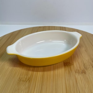 Puede incluir: Un pequeño plato para hornear de forma ovalada con un exterior amarillo y un interior blanco. El plato tiene dos asas a los lados.