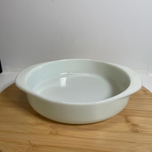 May include: A white, oval-shaped ceramic baking dish with two handles. The dish has a smooth, glossy finish and is set on a wooden surface. The dish is empty and ready for use.