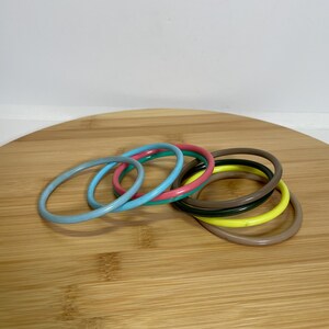 Puede incluir: Una colección de pulseras coloridas en varios tonos, incluyendo azul claro, turquesa, rosa, verde, amarillo y marrón. Las pulseras están dispuestas sobre una superficie de madera, mostrando su diseño liso y redondeado. Estos accesorios son perfectos para añadir un toque de color a cualquier atuendo.