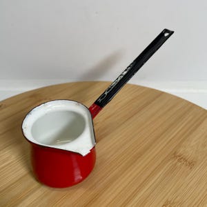 Könnte beinhalten: Ein kleiner, roter Emaille-Kochtopf mit weißem Innenraum und einem langen, schwarzen Griff. Der Griff hat ein dekoratives Design. Der Kochtopf steht auf einer Holzoberfläche.