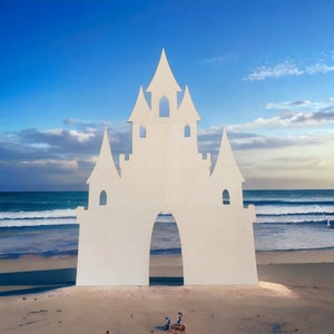 Può includere: Un ritaglio di castello di cartone bianco su una spiaggia sabbiosa con onde blu dell'oceano sullo sfondo.