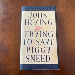 May include: A hardcover book titled "Trying to Save Piggy Sneed" by John Irving. The book cover features the author's name in large, dark blue letters, with the title below. A pink pig illustration is above the title. The book has a light yellow background.